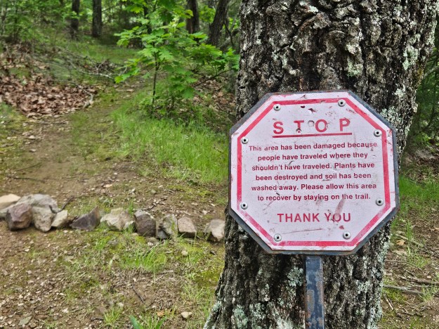 A STOP sign on a tree on a trail that reads "STOP. This area has been damaged because people have traveled where they shouldn't have traveled. Plants have been destroyed and soil has been washed away. Please allow this area to recover by staying on the trail." 