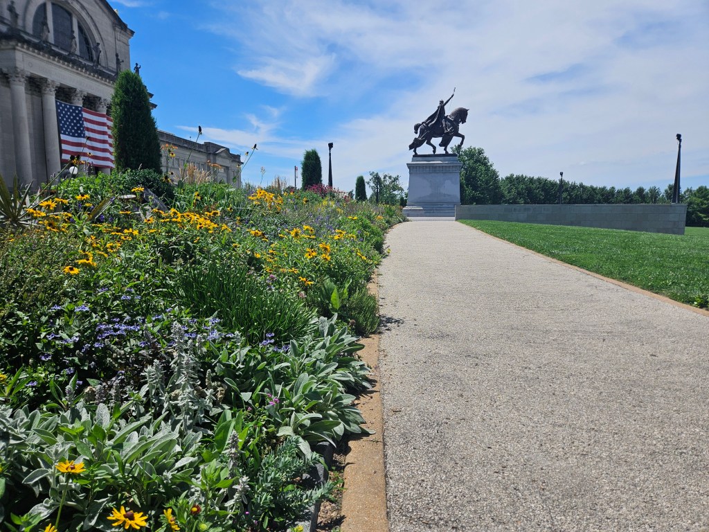 A stature of Saint Louis in front of a building with stone columns and an American Flag. A garden with yellow flowers separates the statue and the building.